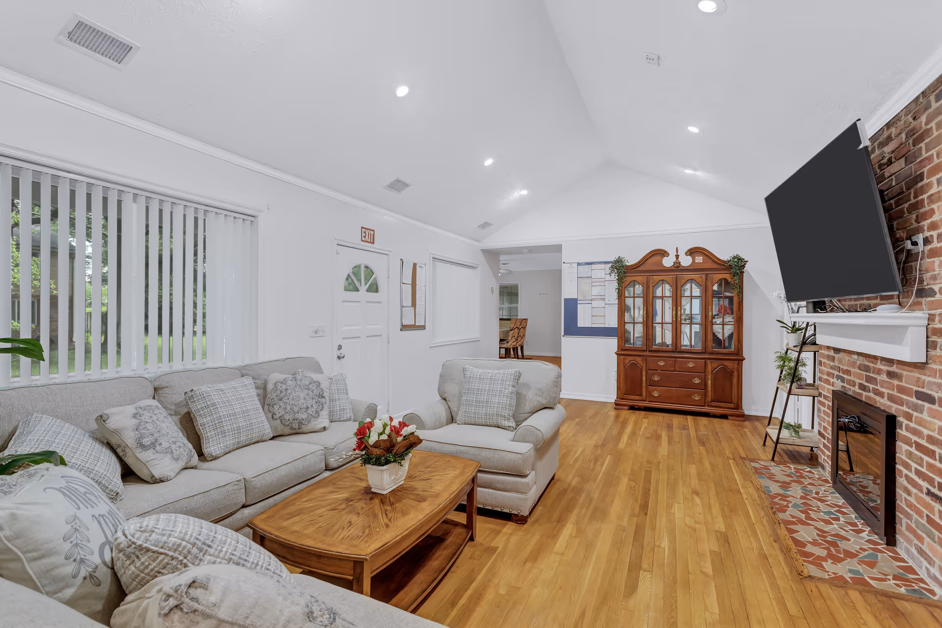 Bright living room with sofas and pillows, a wooden coffee table, a fireplace with a mounted TV, and a china cabinet.