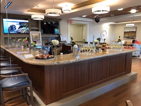 Interior view of a senior living facility's common area featuring a large granite countertop island with bar stools, decorated with flowers, trays of cupcakes, champagne bottles in ice buckets, and glasses filled with champagne. Two flat-screen TVs are mounted on the wall, and a few people are seated in the background on comfortable chairs.