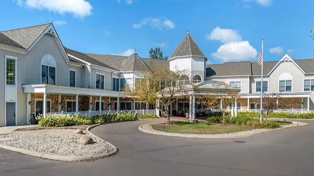 Front exterior of a two-story senior living building with a covered circular drive, landscaped island, and American flag.