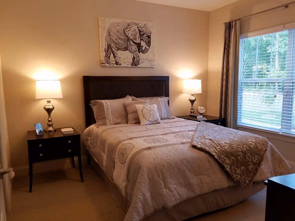 A cozy bedroom with a neatly made bed featuring beige and cream bedding. There are two matching bedside tables with lamps on each side of the bed. A framed picture of an elephant hangs on the wall above the headboard. A window with blinds and curtains lets in natural light on the right side of the room.