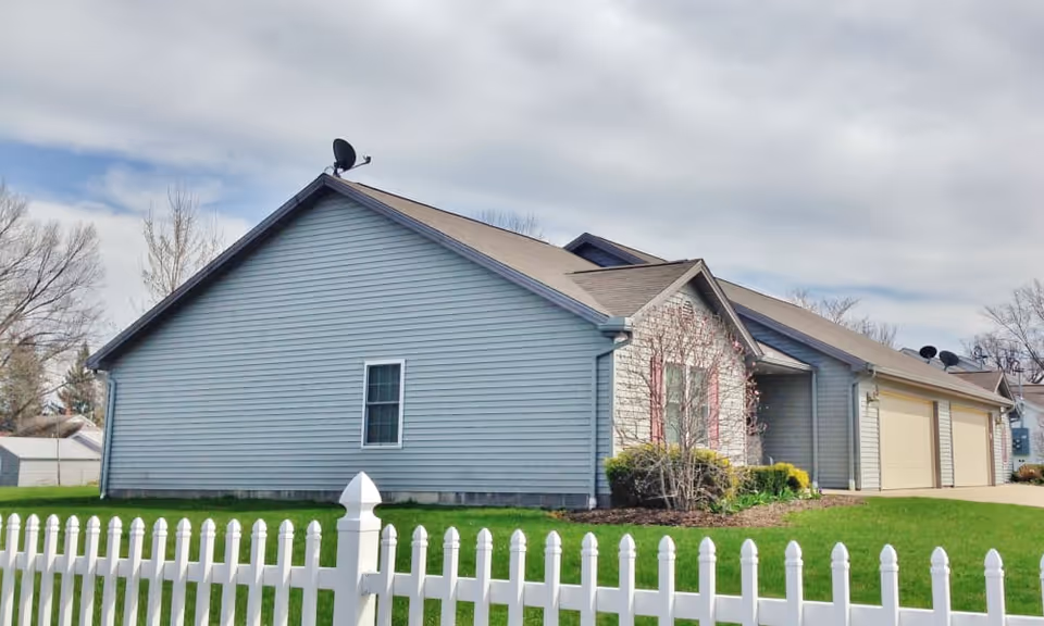 Exterior view of a single-story residential building with light blue siding, a brown shingled roof, and a white picket fence in the foreground. There are some shrubs and a small tree near the entrance, and the sky is partly cloudy.
