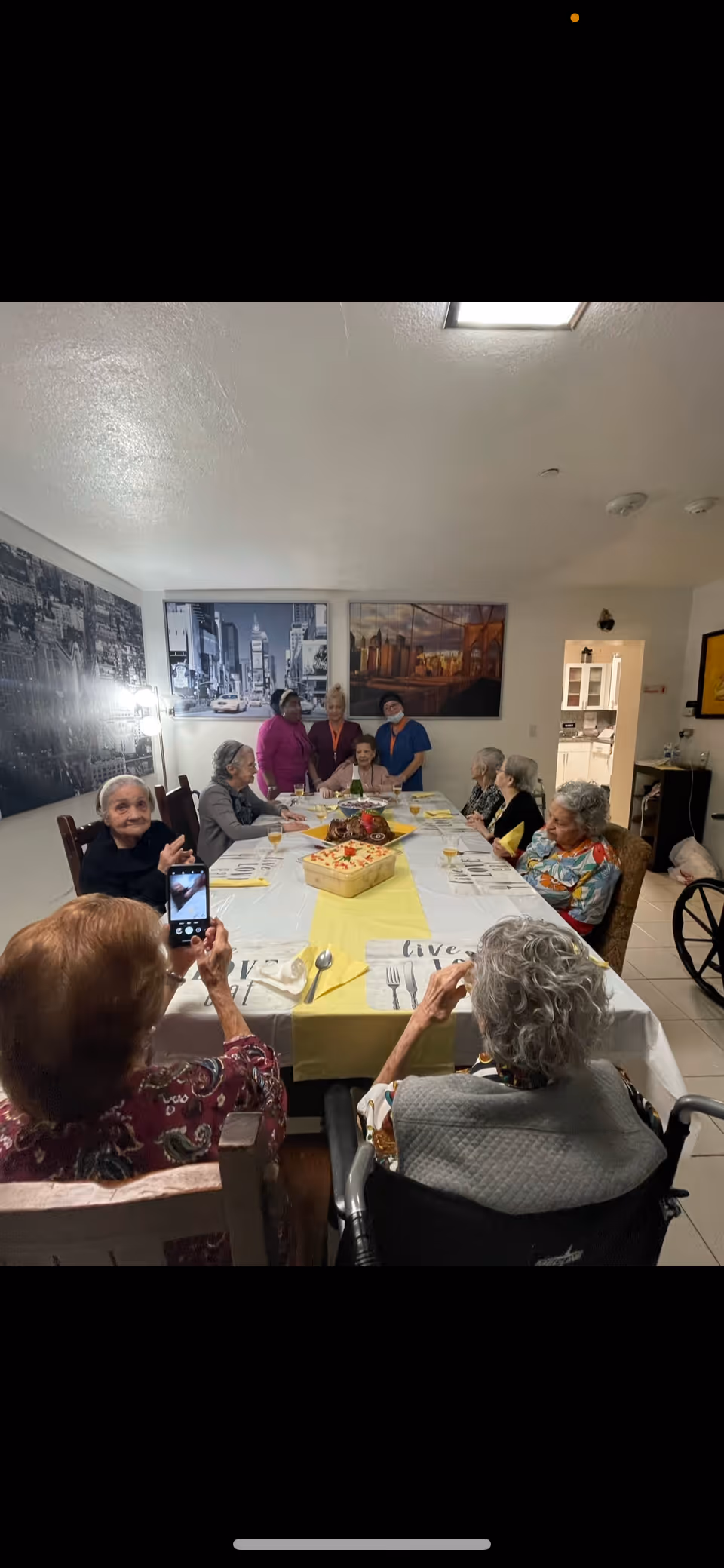 A group of elderly people gathered around a long dining table set with plates, glasses, and a centerpiece. Some are seated while others stand at the far end of the table. The room has framed cityscape artwork on the walls and a bright overhead light. One person is taking a photo with a smartphone.