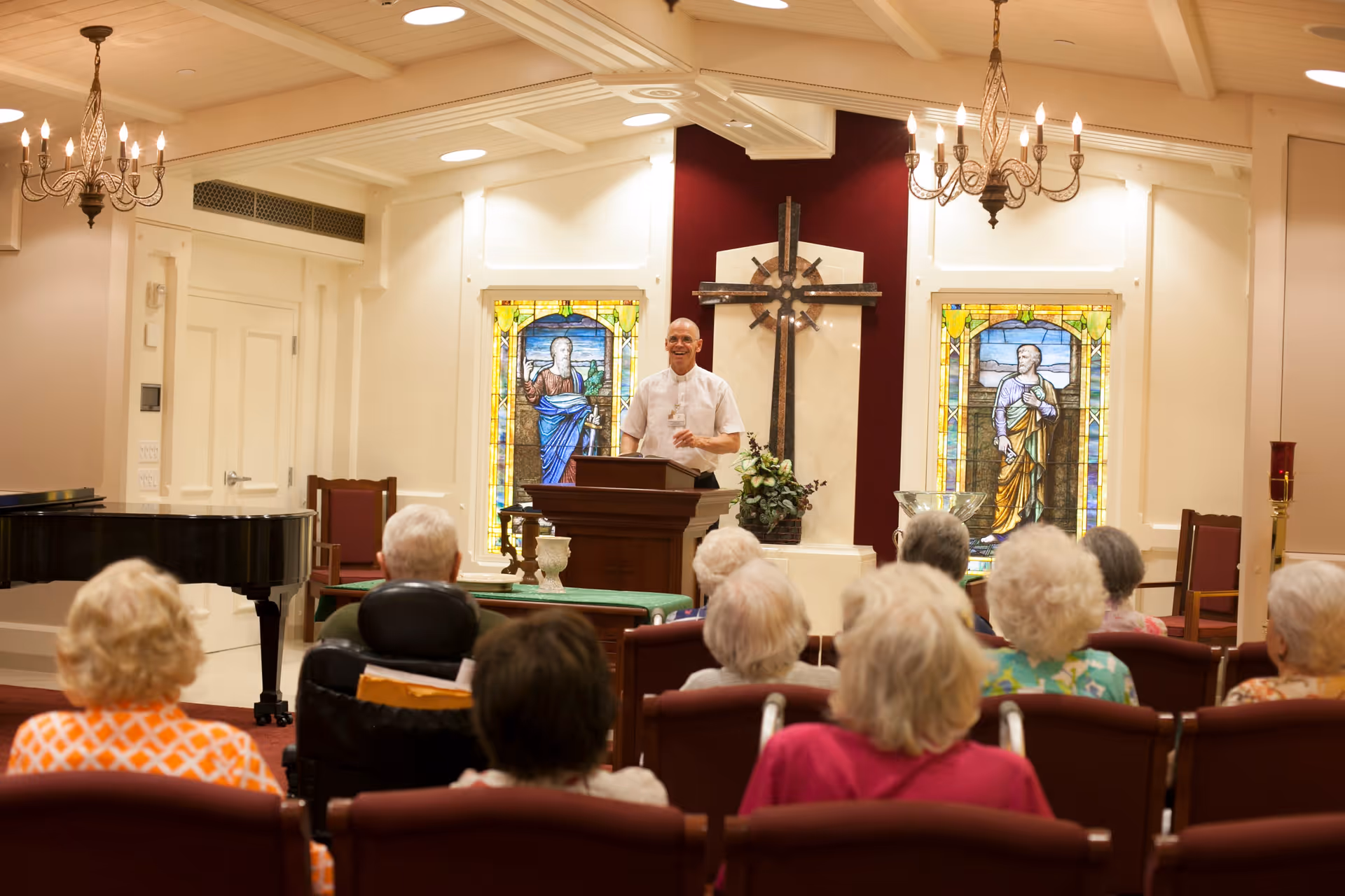 A clergyman leads a service at a chapel pulpit while senior residents sit in rows facing him beneath chandeliers and stained glass windows.