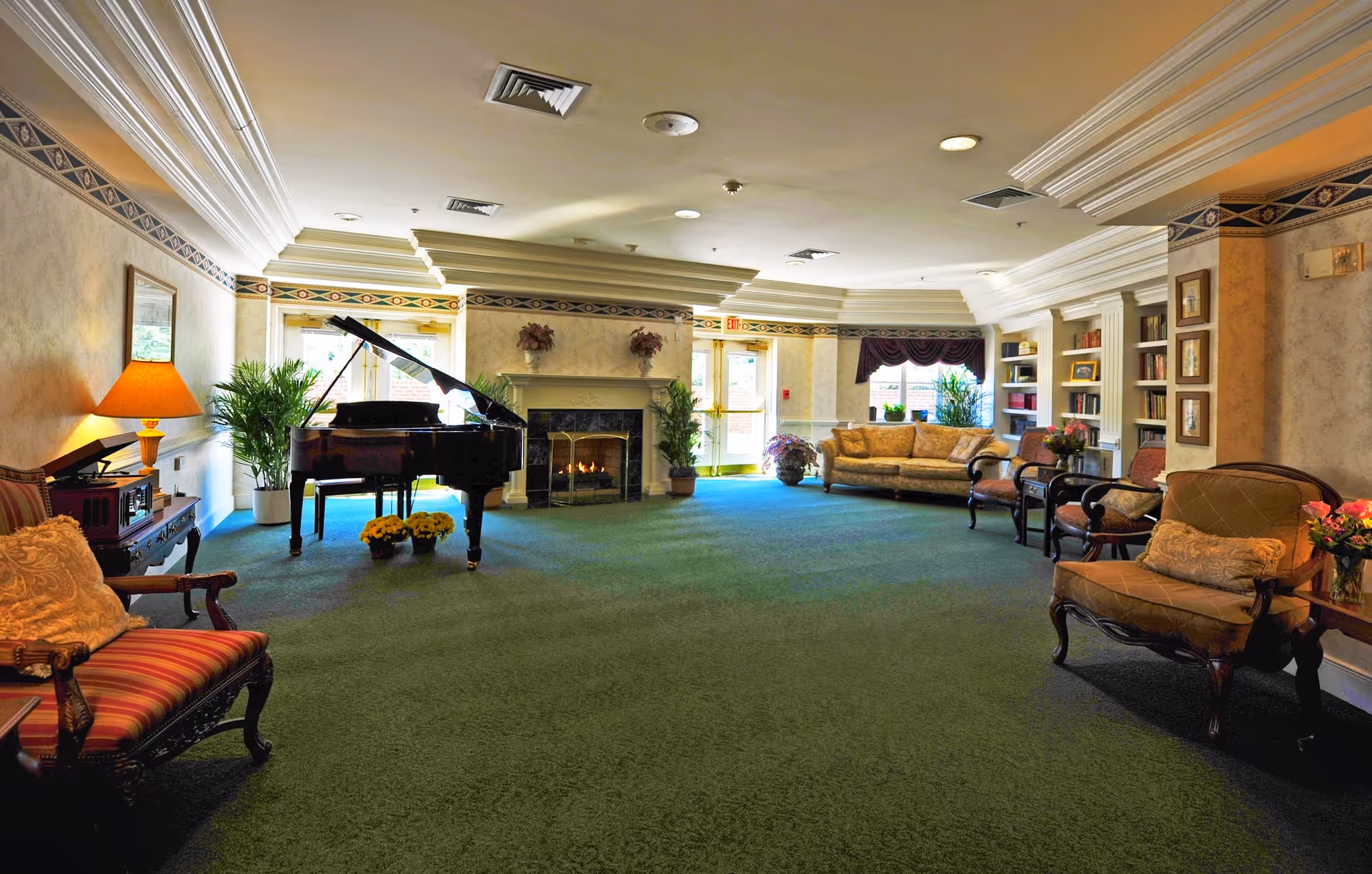 A spacious and elegant living room area with green carpet, a grand piano near a fireplace, several upholstered chairs and sofas, potted plants, a table lamp, and built-in bookshelves. The room has decorative crown molding and patterned wallpaper, with natural light coming through glass doors and windows.