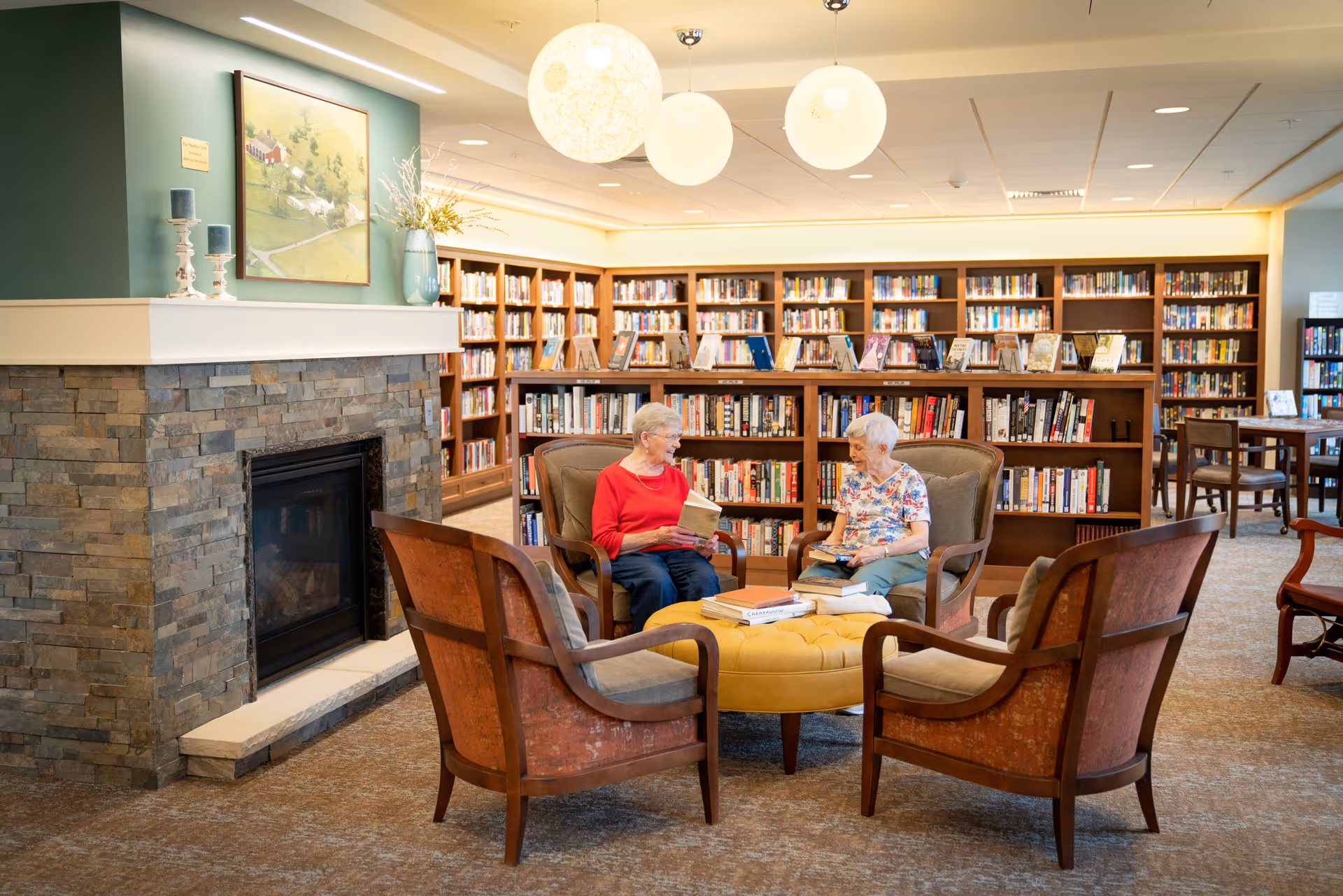 Two elderly women seated in armchairs read and talk in a cozy library-style common room with bookshelves, a fireplace, and round overhead lights.