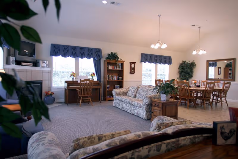 A spacious living room area with floral patterned sofas, a wooden bookshelf, a piano with a chair, and a dining table with chairs. The room has large windows with blue valances, a wall clock, indoor plants, and hanging light fixtures.