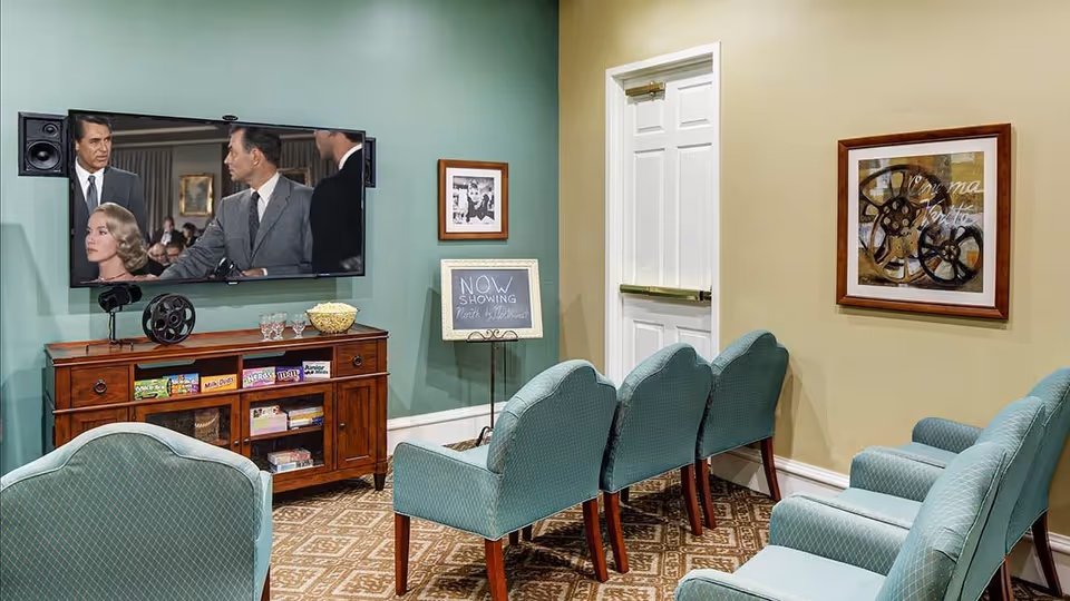 A small movie viewing room with six teal upholstered chairs arranged in two rows facing a wall-mounted flat-screen TV playing a black-and-white film. Below the TV is a wooden cabinet holding snacks and DVDs. A small chalkboard sign reads 'NOW SHOWING North by Northwest.' The walls are painted green and beige, with framed artwork and a white door visible.