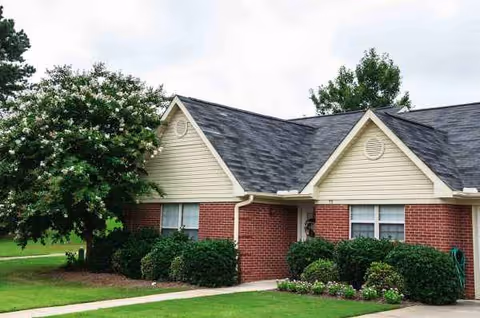 A single-story brick senior living building with beige siding, shrubs, and a manicured front lawn.