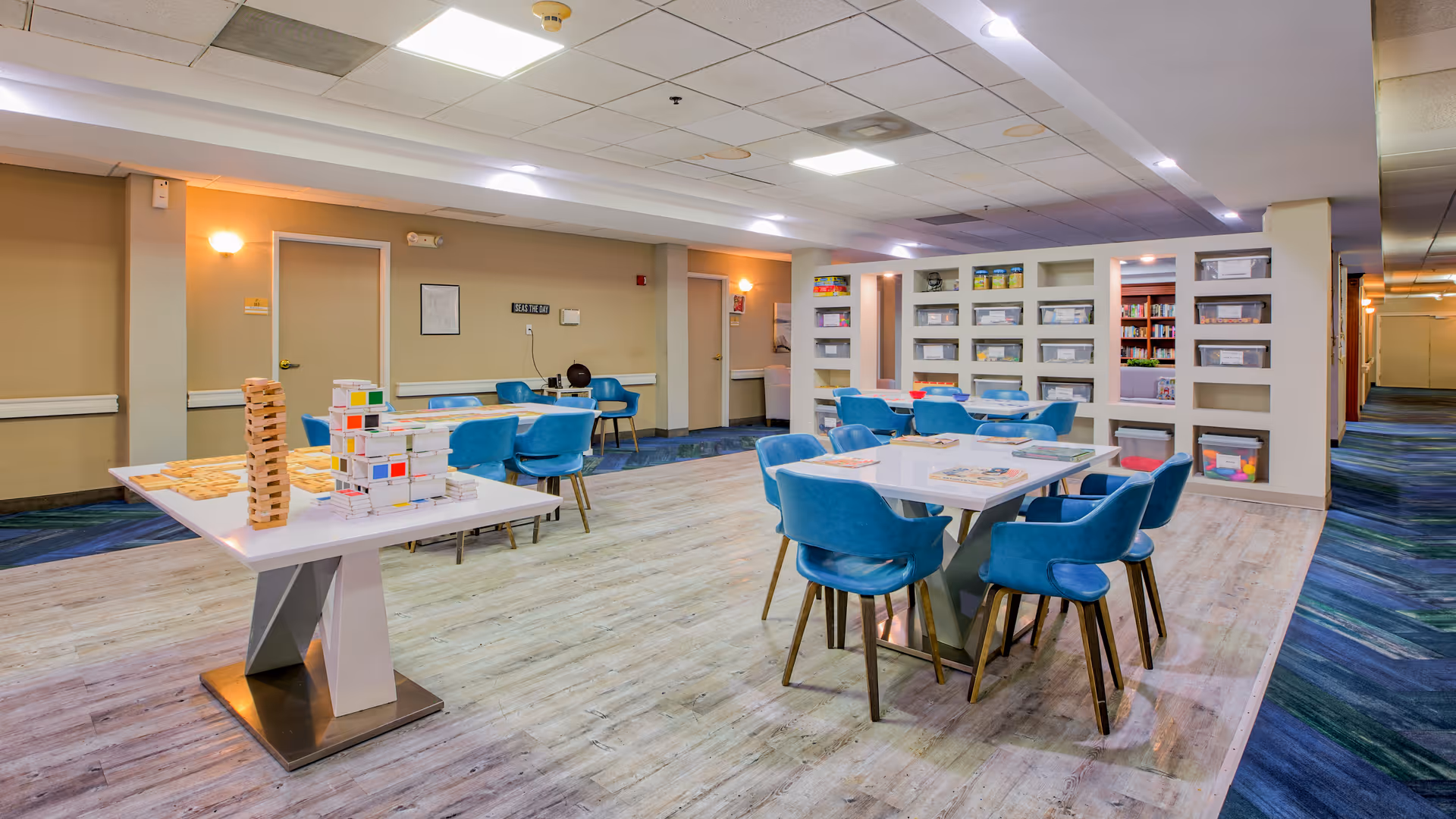 Activity room with several white tables and blue chairs, shelves of games and books, and game sets on a central table.