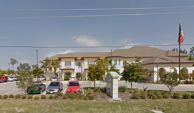 Front exterior of The Woodlands Assisted Living Center building with parked cars, landscaping, and an American flag.
