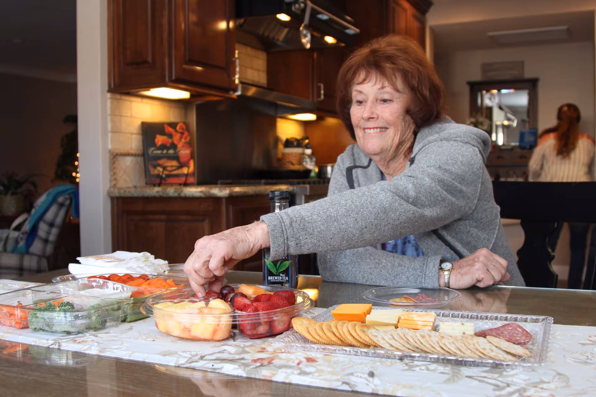 An elderly woman in a gray sweater is sitting at a dining table reaching for grapes from a platter of assorted fruits, cheeses, crackers, and vegetables in a warmly lit kitchen setting.