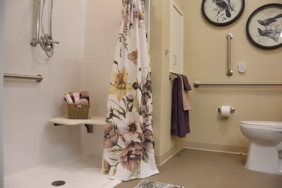 A bathroom with a walk-in shower featuring a floral shower curtain and a built-in fold-down seat holding a basket with toiletries. The room has beige walls with two framed bird illustrations, grab bars near the toilet, a toilet paper holder, and towels hanging on a wall-mounted rack.