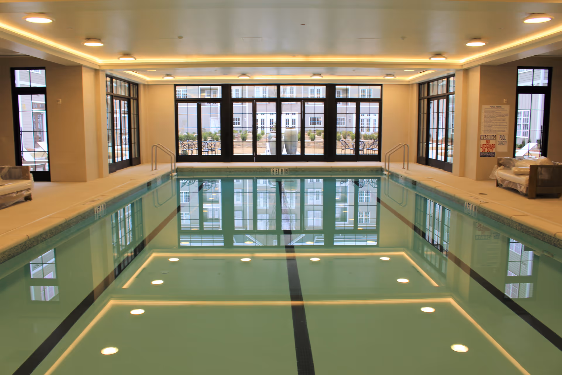 Indoor swimming pool with clear water, surrounded by beige tiled flooring and large windows that let in natural light. There are metal handrails on both sides of the pool and seating areas with covered furniture along the right side. Outside the windows, there is a patio area with tables and chairs.
