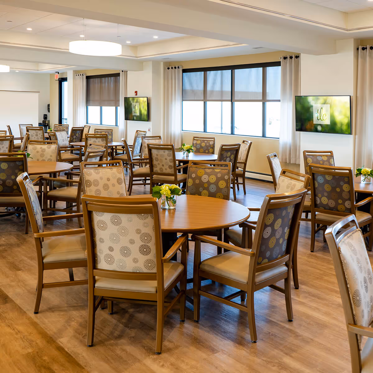 Bright communal dining room with multiple round tables and patterned chairs arranged on wood floors.