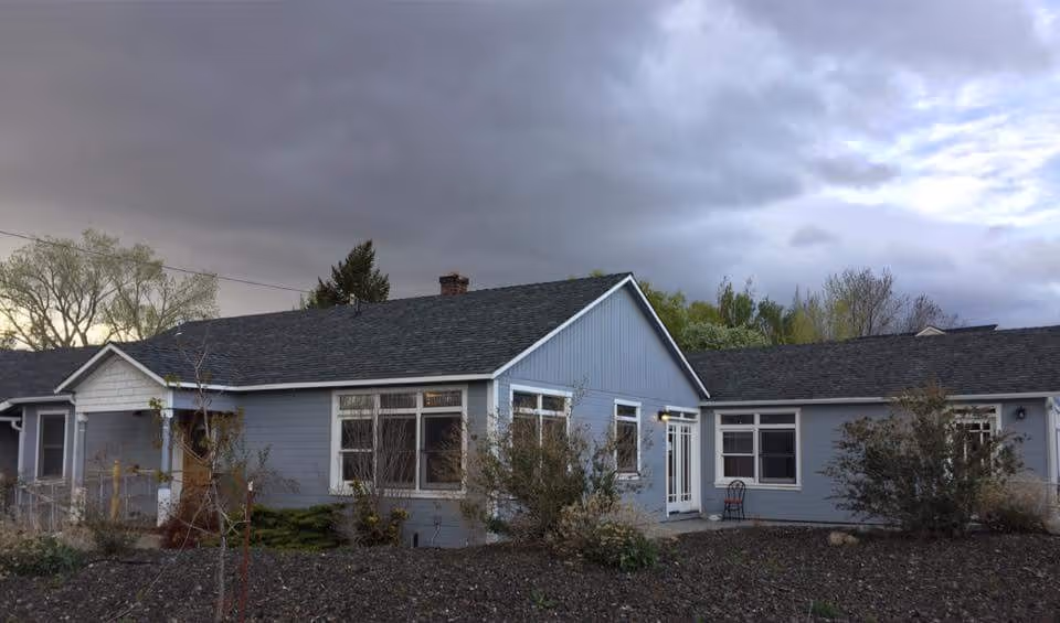 Exterior view of a single-story residential care facility building with light blue siding and a dark shingled roof under a cloudy sky. There are several windows and a white door with glass panels. Some bushes and small trees are planted around the building.