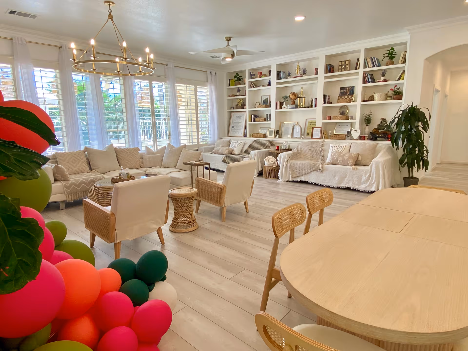 A bright and spacious living room with large windows covered by white sheer curtains and plantation shutters. The room features a large beige sectional sofa, two armchairs with wooden arms, a round wicker side table, and a wooden dining table with chairs. Built-in white shelves filled with books, plants, and decorative items line the back wall. A modern chandelier and ceiling fan hang from the ceiling. Colorful balloon decorations are visible in the foreground on the left side.