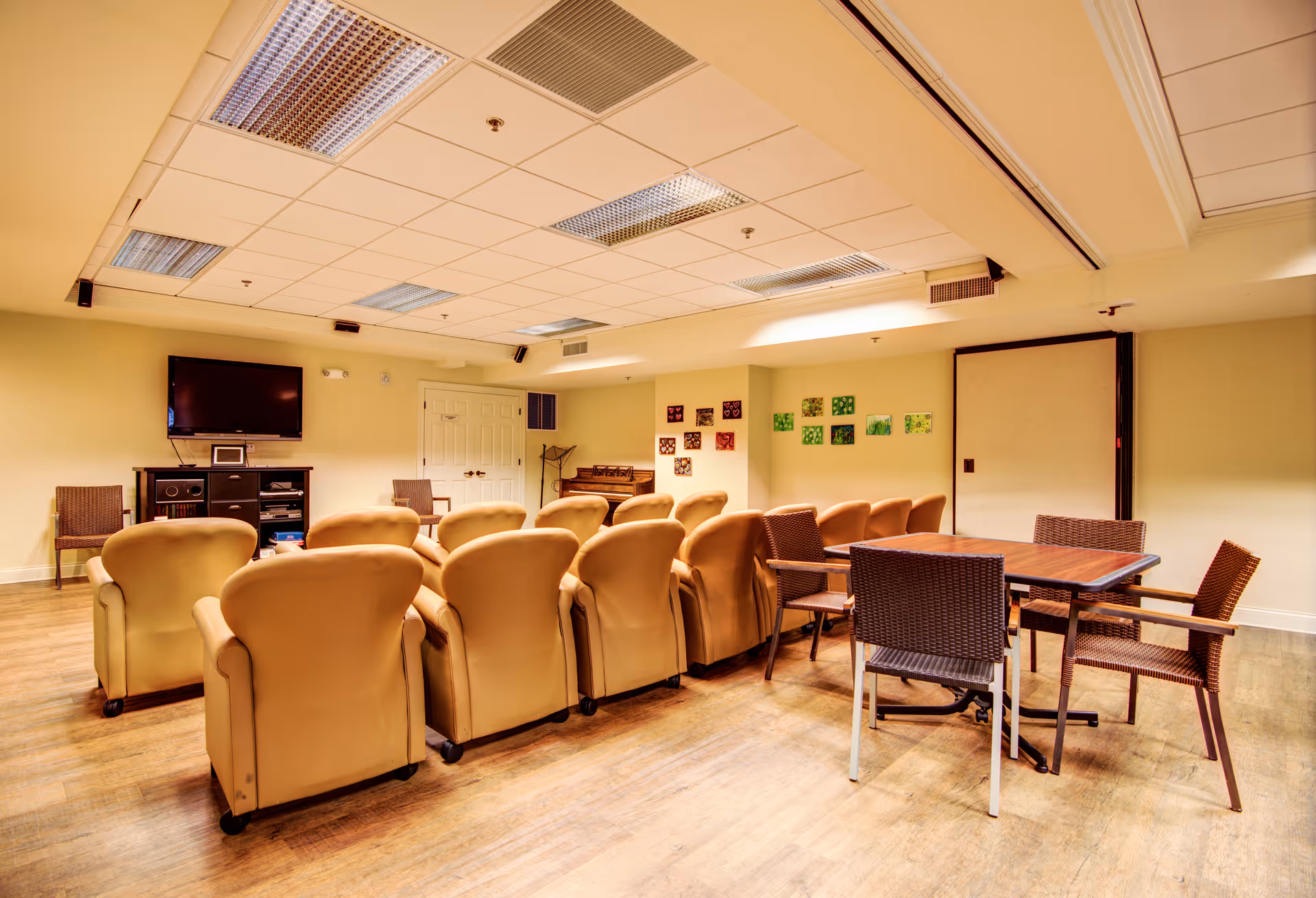 A cozy common room with two rows of beige armchairs facing a wall-mounted flat screen TV. To the right, there is a wooden table with four wicker chairs. The room has a light yellow wall with small colorful square artworks and a piano in the corner. The floor is wooden, and the ceiling has fluorescent lights.