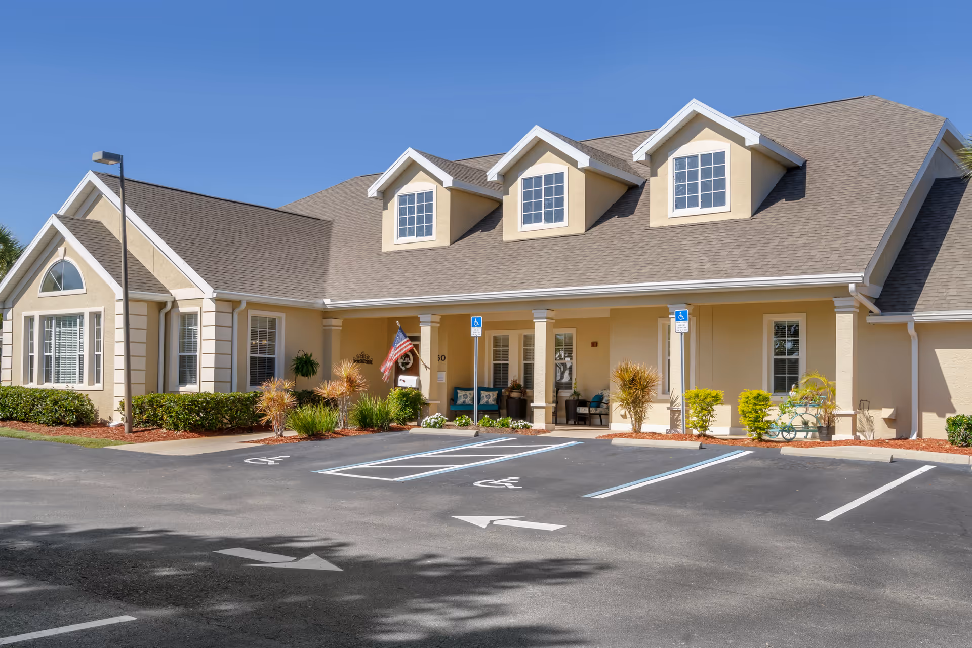 Front exterior of a beige senior living building with a covered entrance, three dormer windows, accessible parking spaces, and an American flag.