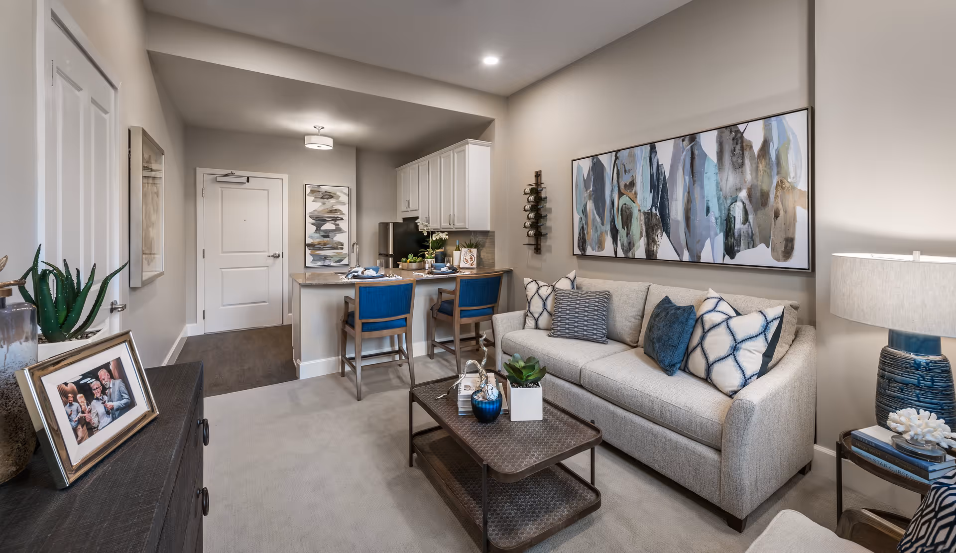 A cozy living area in an assisted living facility featuring a beige sofa with patterned cushions, a metal coffee table with decorative items, a side table with a blue lamp and books, and a small kitchen area with a counter and two blue chairs. The walls are decorated with abstract artwork and the space is softly lit with ceiling and table lamps.