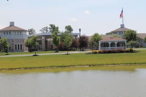 View of a senior living facility named Bristol Village with a large pond in the foreground, a well-maintained lawn, trees, a white gazebo, and buildings with multiple windows and an American flag flying on a flagpole.