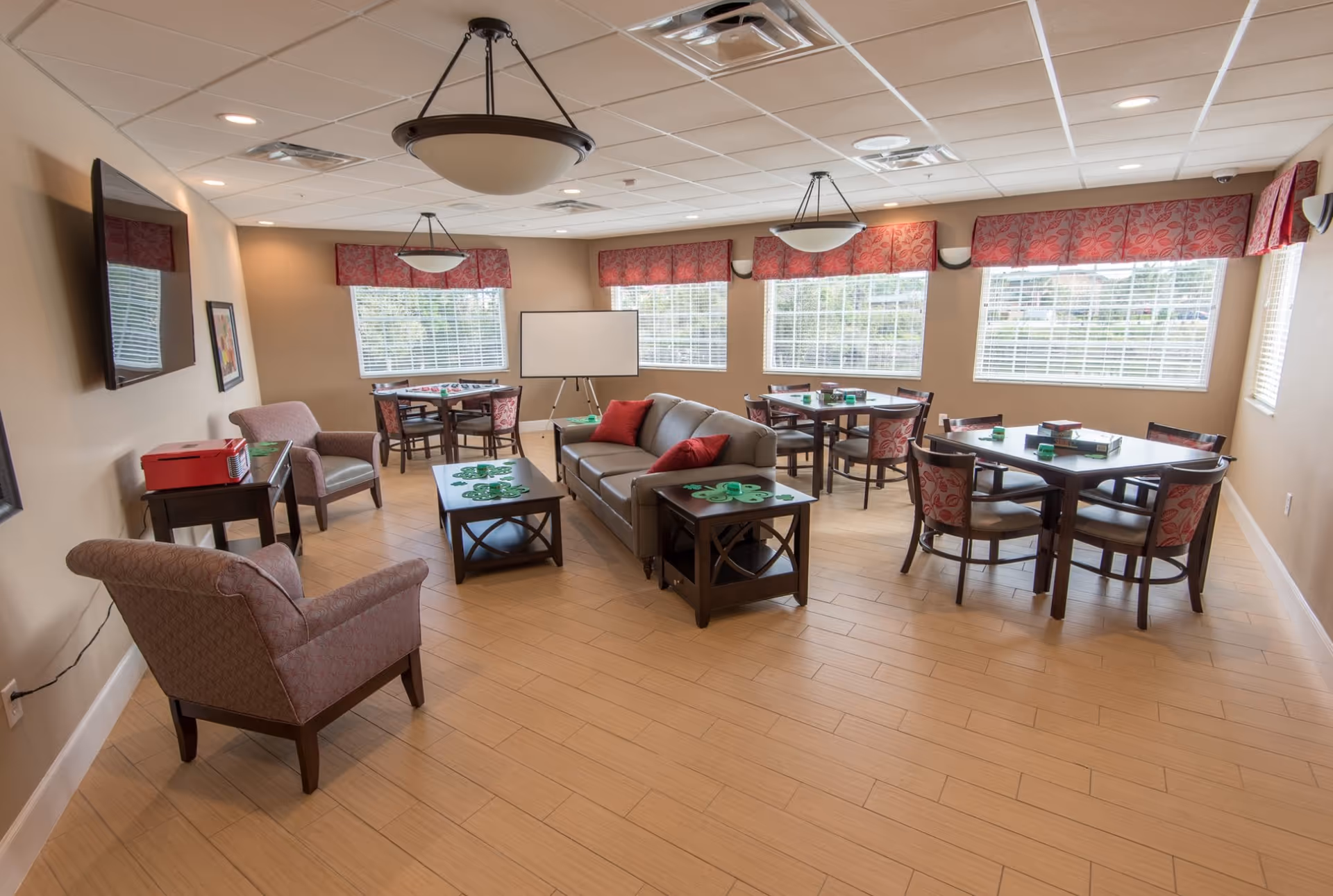 Bright communal activity room with sofas, armchairs, multiple game tables, a wall-mounted TV, and large windows.