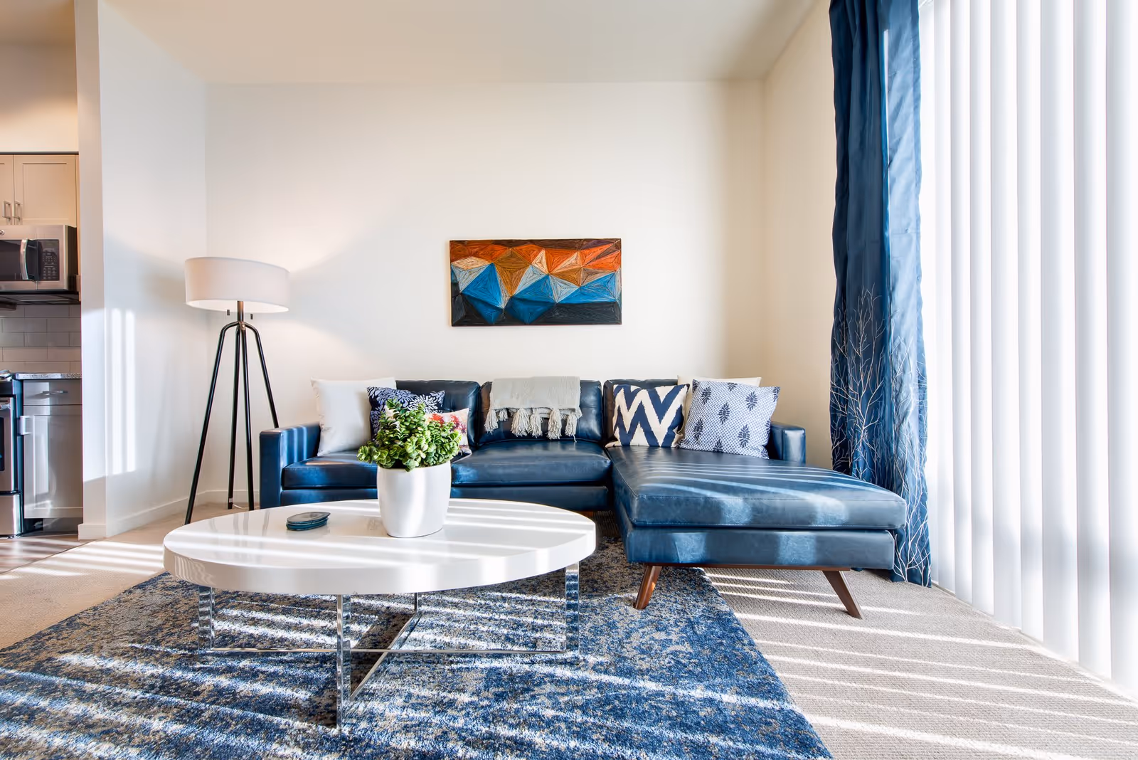 A bright living room with a blue sectional sofa adorned with patterned pillows and a throw blanket. A white oval coffee table with a potted plant sits on a blue patterned rug. A tall floor lamp stands to the left, and a colorful geometric painting hangs on the wall above the sofa. Large vertical blinds cover the window on the right, allowing sunlight to stream in.