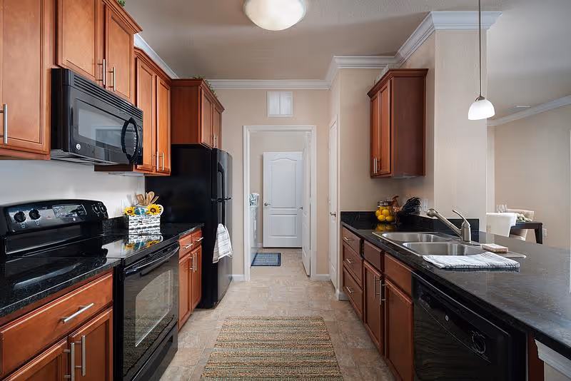 A modern kitchen with dark granite countertops and wooden cabinets. The kitchen features a black stove with an oven, a microwave above it, a black refrigerator, and a double sink with a faucet. There is a small rug on the tiled floor and a doorway leading to another room. The kitchen opens up to a dining area with a table and chairs visible in the background.
