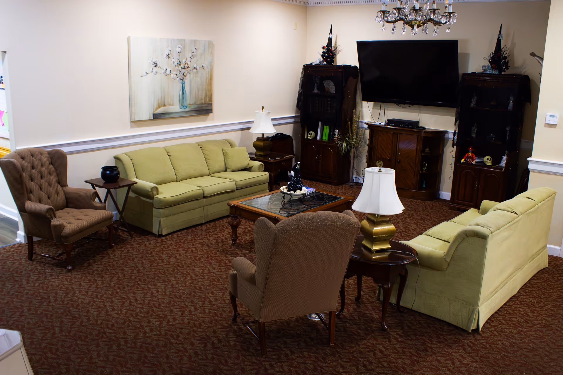 Communal living room with two green sofas, upholstered armchairs, a coffee table, lamps, and a wall-mounted TV with wooden cabinets.