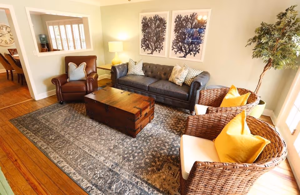 A cozy living room with a dark gray tufted sofa adorned with patterned pillows, a brown leather armchair with a blue pillow, and two wicker chairs with yellow pillows. A wooden chest serves as a coffee table on a large patterned rug. The room has light green walls, two framed botanical prints, a floor lamp, a potted plant near a window, and hardwood floors. A dining area is visible through an open wall space.