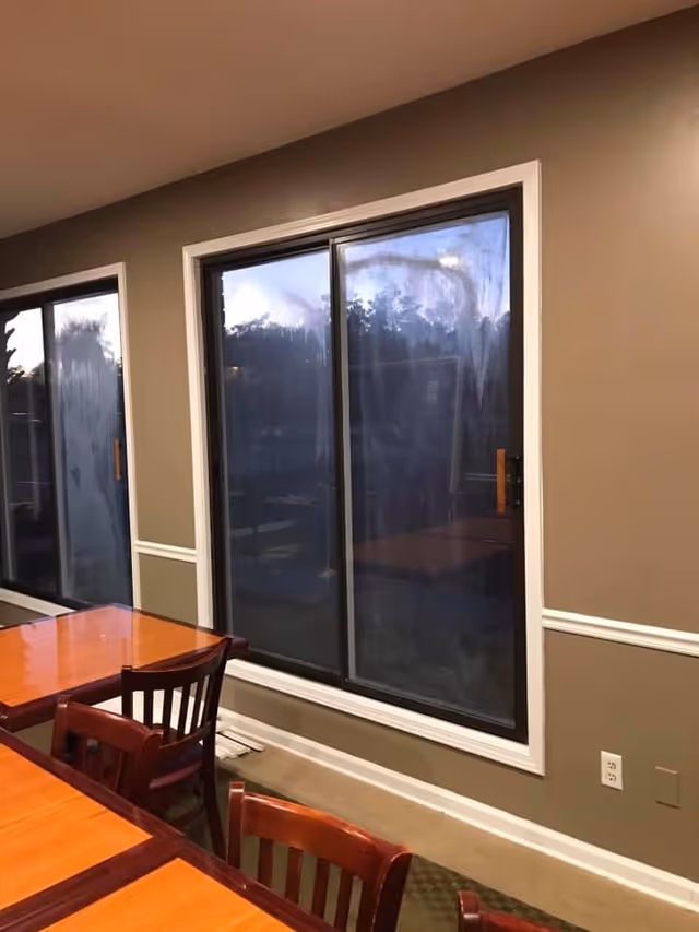 Interior view of a room with beige walls and large sliding glass windows. There are wooden tables and chairs arranged near the windows, and the room has white trim along the walls.