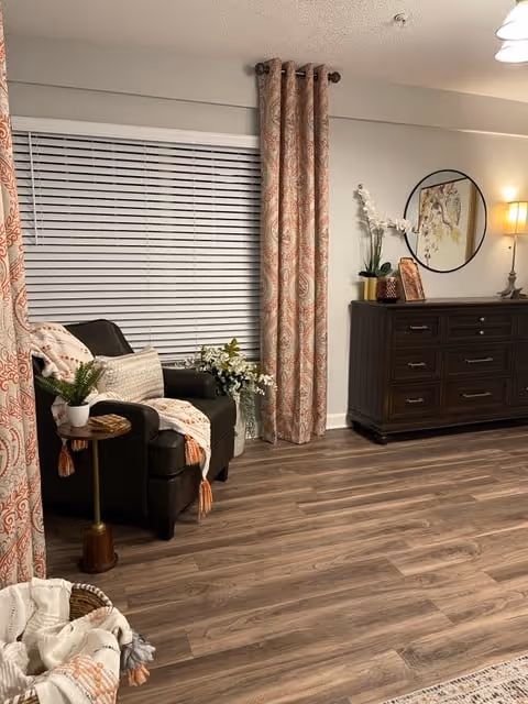 A cozy living room area featuring a dark armchair with a white and orange throw blanket and pillow, a small round side table with a potted plant, floral curtains on a large window with closed blinds, a dark wooden dresser with decorative items including a lamp, a round mirror, and a vase with white flowers, all set on wood flooring.