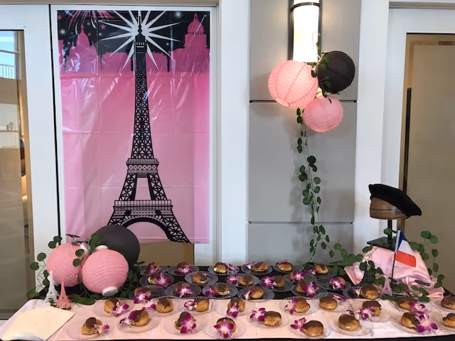 A table decorated with pink and black paper lanterns, purple orchids, and small pastries on plates. Behind the table is a pink backdrop featuring a black silhouette of the Eiffel Tower. A small French flag and a black beret are also part of the decoration.