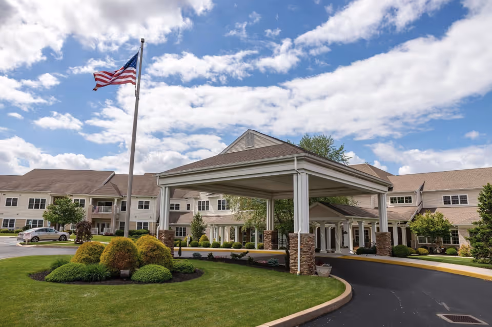 Front entrance of a multi-story senior living building with a covered porte-cochère, landscaped lawn, and an American flag under a partly cloudy sky.