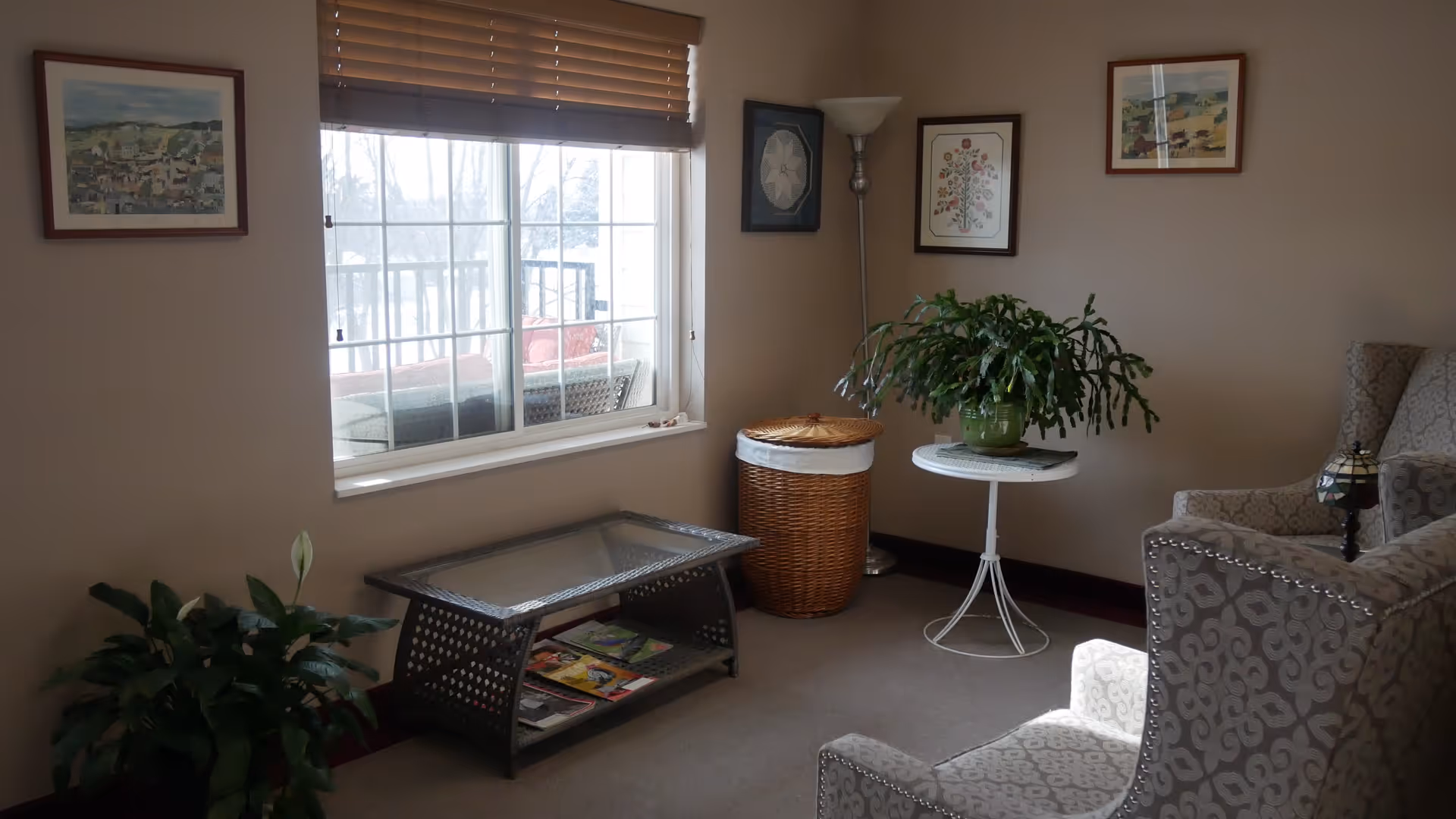 A cozy living room area with a large window covered by wooden blinds. There is a glass-top coffee table with magazines underneath, a wicker basket, a small round white table with a green potted plant, and two patterned armchairs. The walls are decorated with framed artwork, and a tall floor lamp stands in the corner.