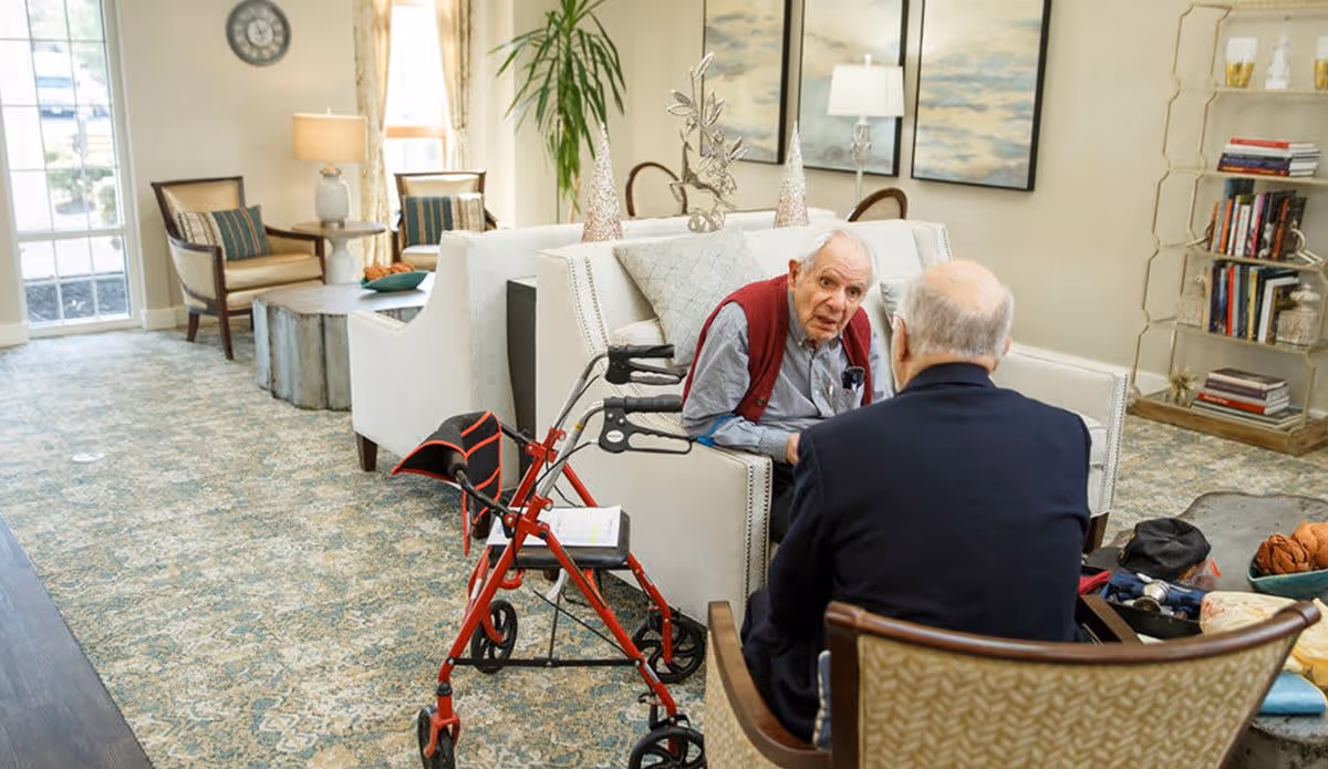 Two elderly men sitting and talking in a well-lit living room with patterned carpet, white sofas, armchairs, a coffee table, and decorative items. One man is seated on a sofa with a red walker beside him, while the other man sits in a chair facing him. The room has large windows, lamps, artwork on the walls, and a bookshelf with books and decor.