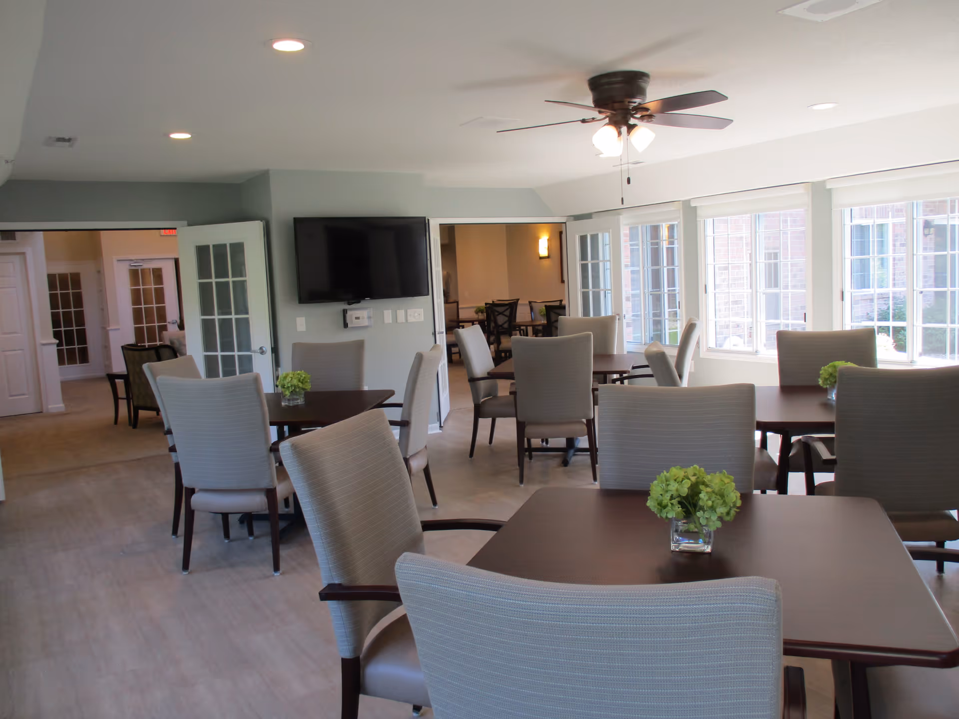 A bright and spacious common area with multiple square tables and cushioned chairs arranged around them. Each table has a small green floral centerpiece. Large windows on one side let in natural light, and a ceiling fan with lights is mounted on the ceiling. A flat-screen TV is mounted on the wall, and there are open doorways leading to other rooms.