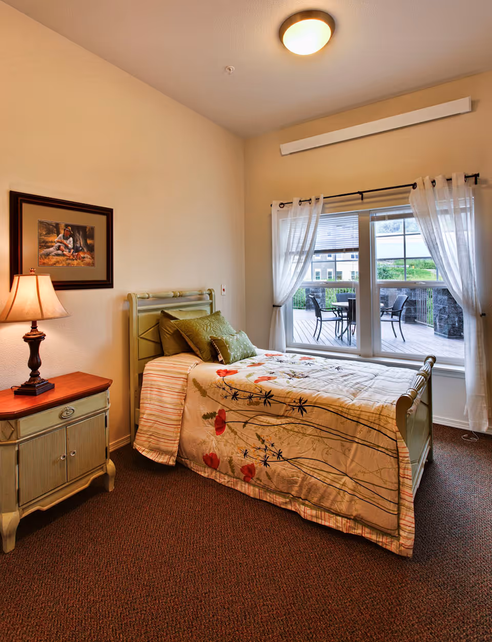A cozy bedroom in a senior living community featuring a single bed with floral bedding and green pillows, a wooden nightstand with a lamp, a framed picture on the wall, and a large window with sheer white curtains overlooking an outdoor patio with chairs and a table.