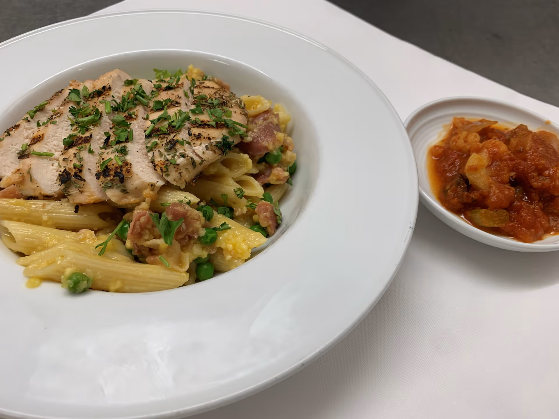 A white plate with grilled sliced chicken breast served over penne pasta mixed with peas and bits of bacon, garnished with chopped herbs. Next to it is a small white bowl containing a chunky tomato-based sauce with vegetables.