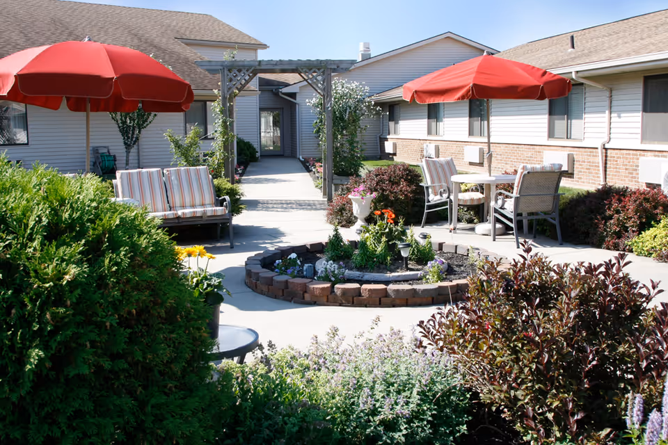 Sunlit courtyard with patio seating, red umbrellas, a central brick-bordered flowerbed and shrubs in front of a single-story building.