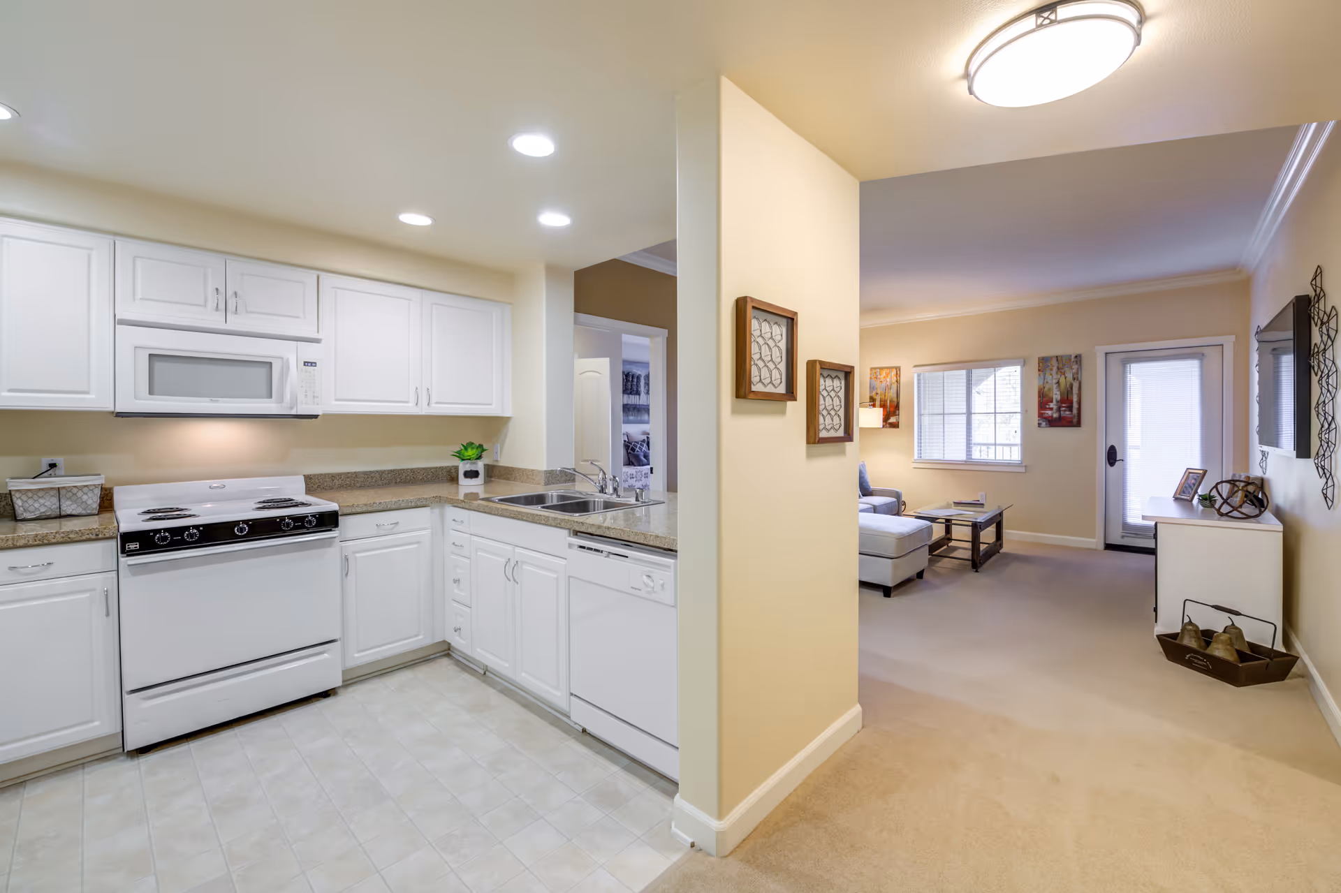 Interior view of a senior living facility showing a kitchen with white cabinets, a stove, microwave, and dishwasher on the left side, and a living room area with a sofa, coffee table, wall-mounted TV, and a door leading outside on the right side. The walls are painted light beige and the floor is tiled in the kitchen and carpeted in the living room.