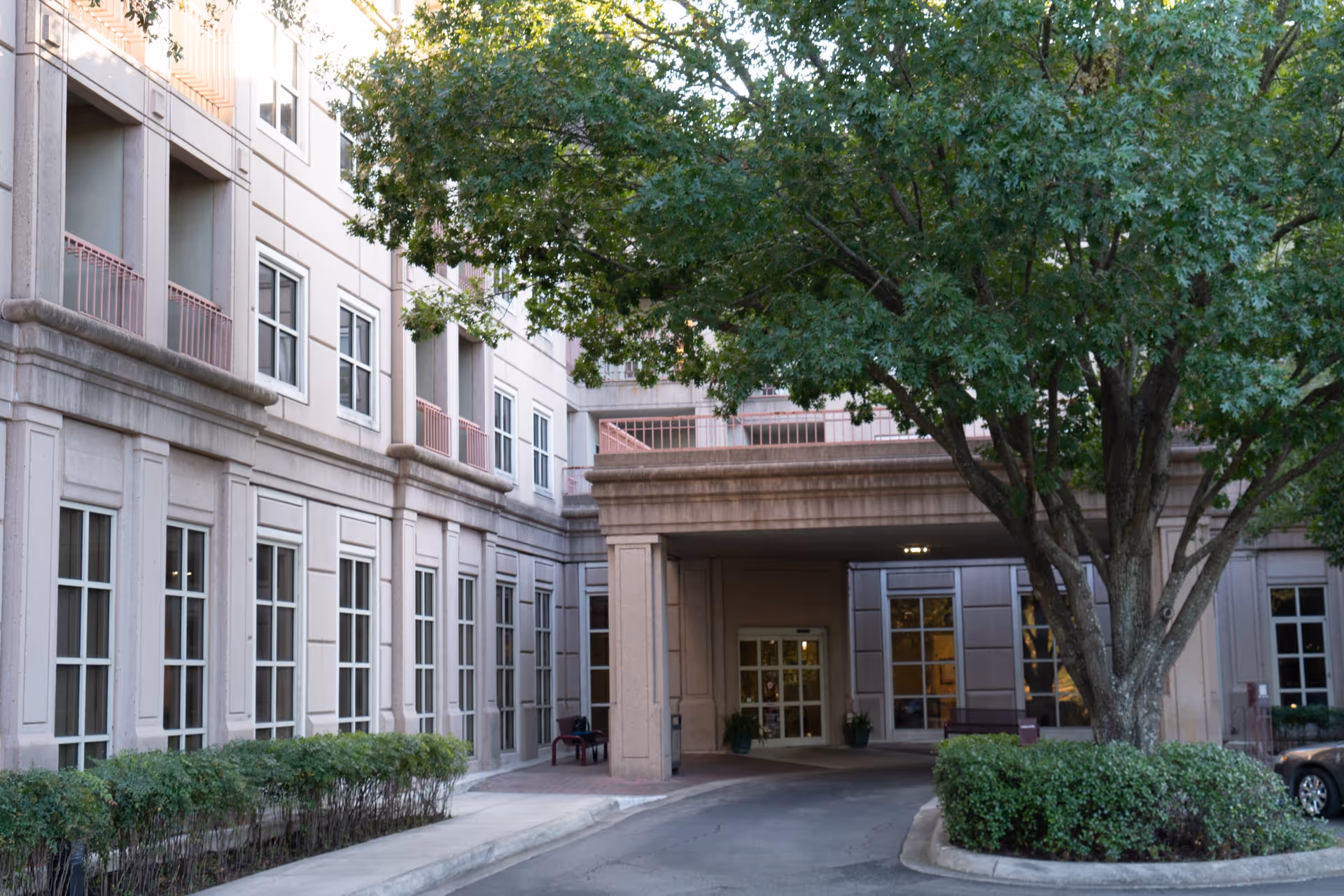 Exterior view of a multi-story healthcare facility building with large windows, a covered entrance driveway, and a large tree with green foliage in front. There are shrubs lining the walkway and a parked car partially visible on the right side.