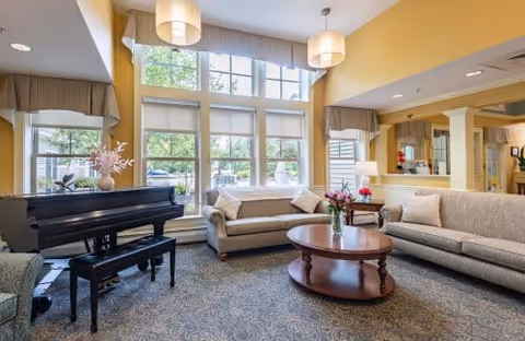 A bright and spacious living room with large windows letting in natural light. The room features two beige sofas with cushions, a round wooden coffee table with a vase of flowers, a black grand piano with a matching bench, and soft yellow walls. The carpet has a subtle pattern, and there are pendant lights hanging from the ceiling.