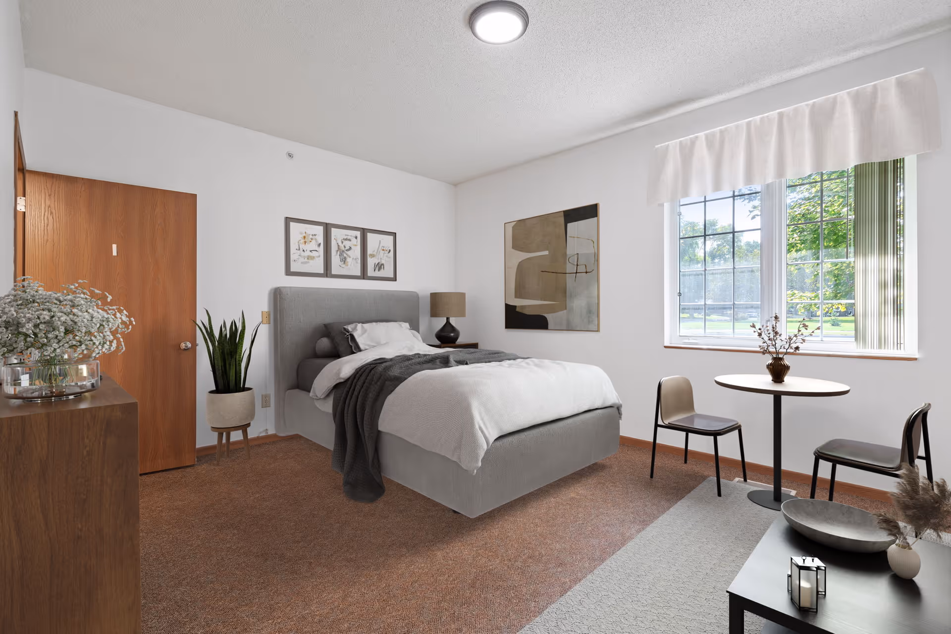 A cozy bedroom with a gray upholstered bed made with white and gray bedding. There is a wooden door on the left wall, a dresser with a vase of flowers, a potted plant, and a bedside table with a lamp. On the right side, there is a round table with two chairs near a window with white curtains, letting in natural light. The walls are white and decorated with framed artwork.