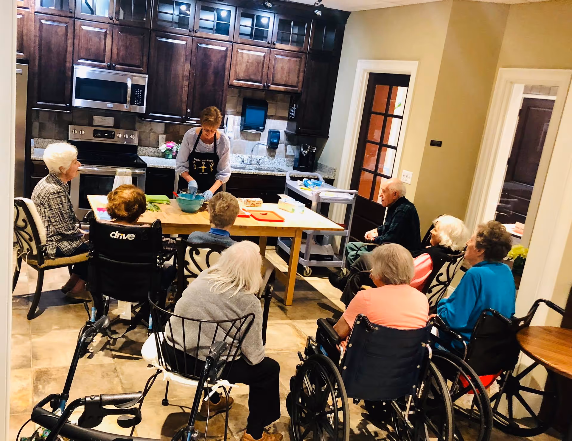 A group of elderly individuals, some in wheelchairs, seated around a kitchen island watching a woman preparing food in a kitchen with dark wooden cabinets and stainless steel appliances.