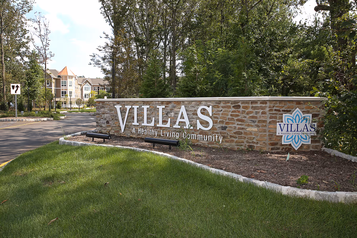 Stone entrance sign reading "VILLAS A Healthy Living Community" with landscaping and the facility buildings visible behind trees.