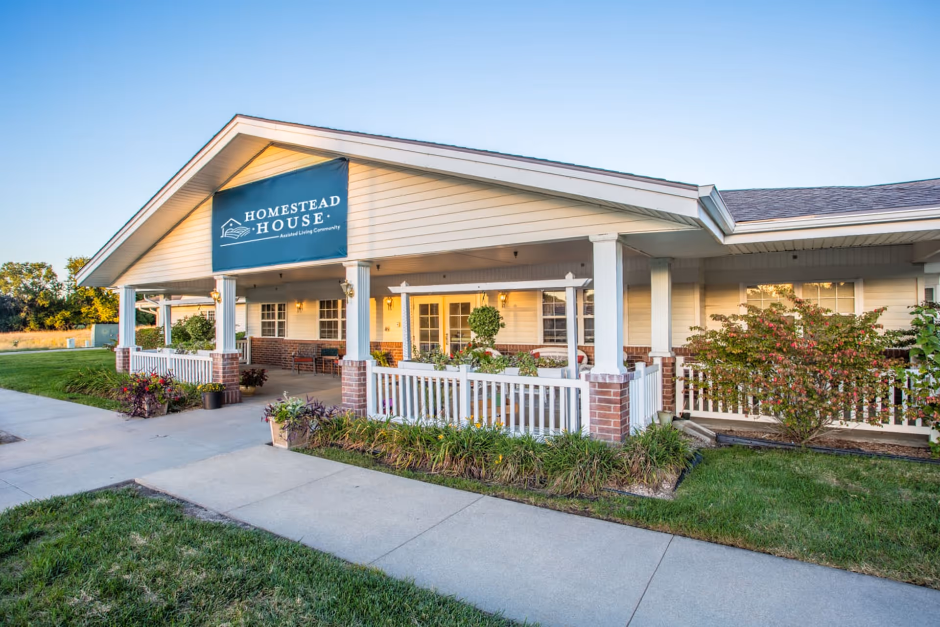 Exterior view of Homestead House, a single-story assisted living community building with a covered porch, white railings, brick pillars, and surrounding greenery under a clear sky.