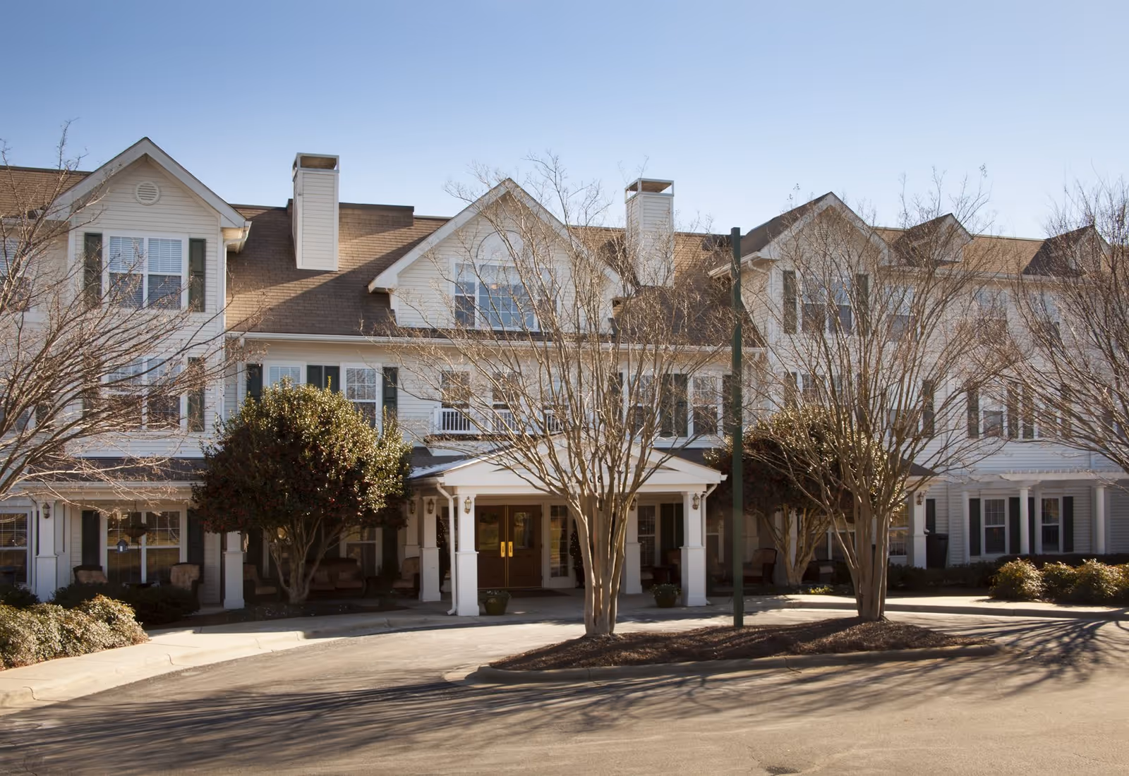 Front exterior of a multi-story senior living facility with a covered entrance, trees, and a circular driveway.