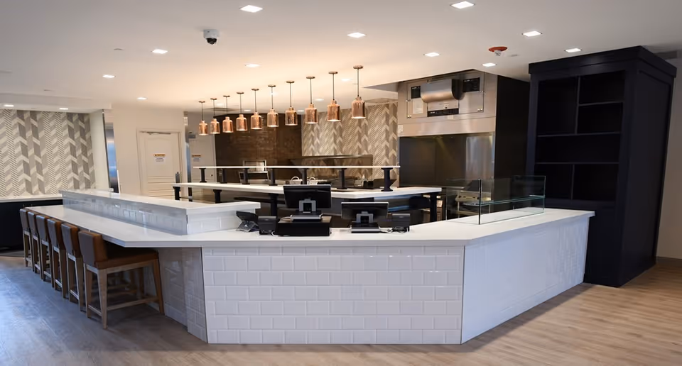 Modern serving counter and open kitchen with bar seating and pendant lights in a senior living dining area.