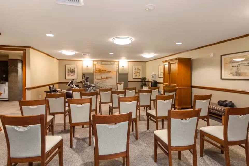 A room set up with multiple wooden chairs with white cushions arranged in rows facing a wall with framed artwork. The room has beige walls with wooden trim, carpeted floor, ceiling lights, and exercise equipment in the back corner.