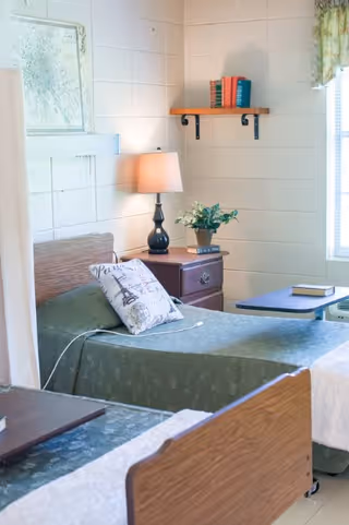 Two single beds in a bright senior living bedroom with a nightstand, lamp, small shelf of books, and a window.